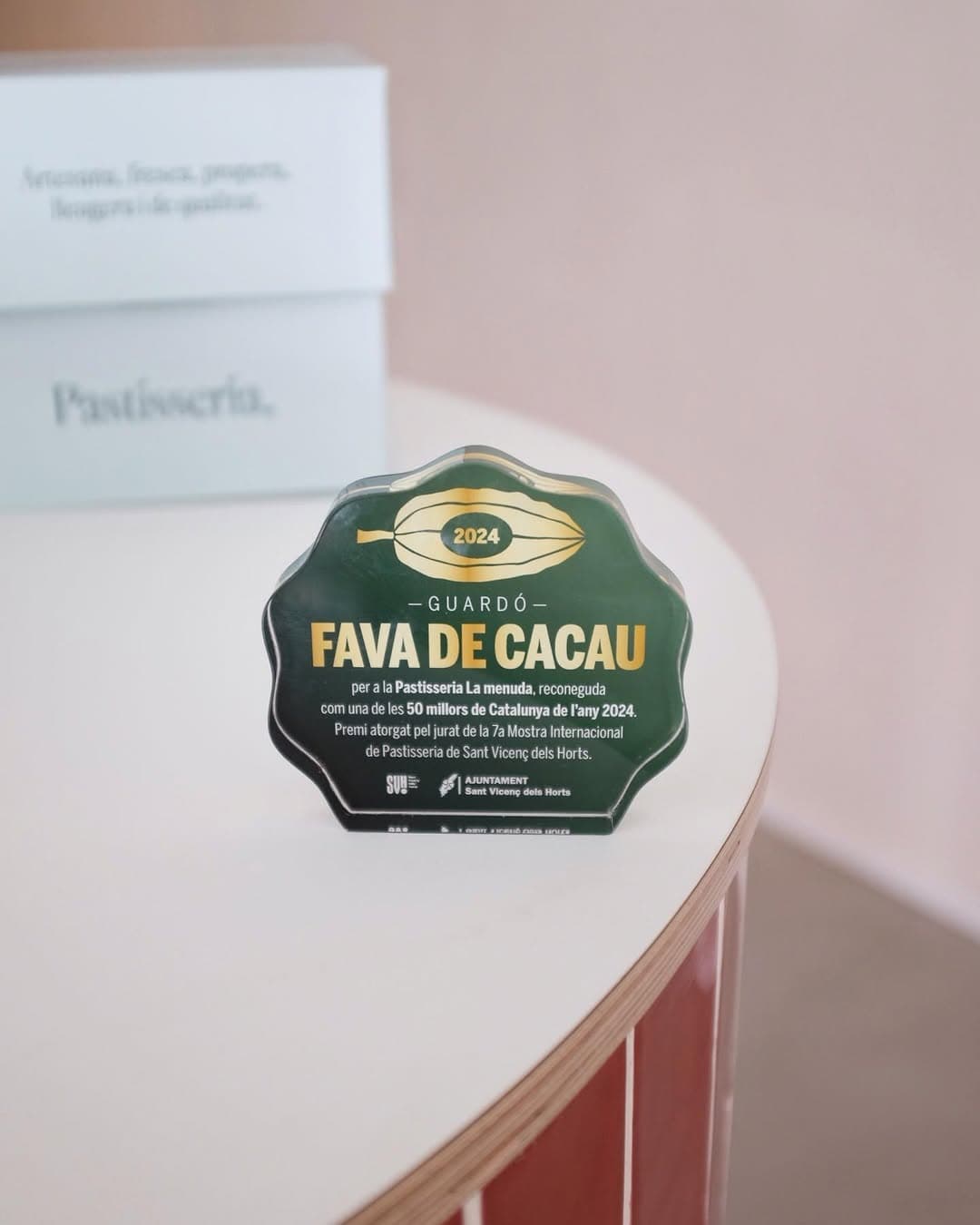Es mostra el trofeu de la fava de cacau de 2024, forma ovalada i de cristall, de color verd amb lletres de color daurat
