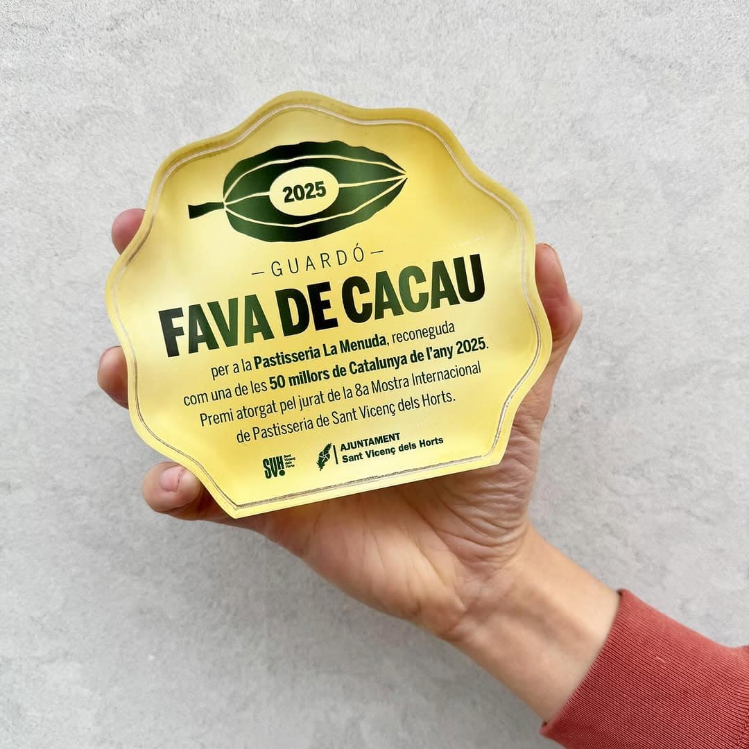 Es mostra el trofeu de la fava de cacau de 2025, forma ovalada i de cristall, de color groc amb lletres de color verd fosc